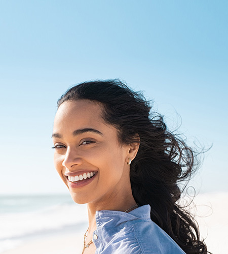 The image depicts a woman with long hair, smiling at the camera, standing on a beach with clear skies and light blue water in the background.