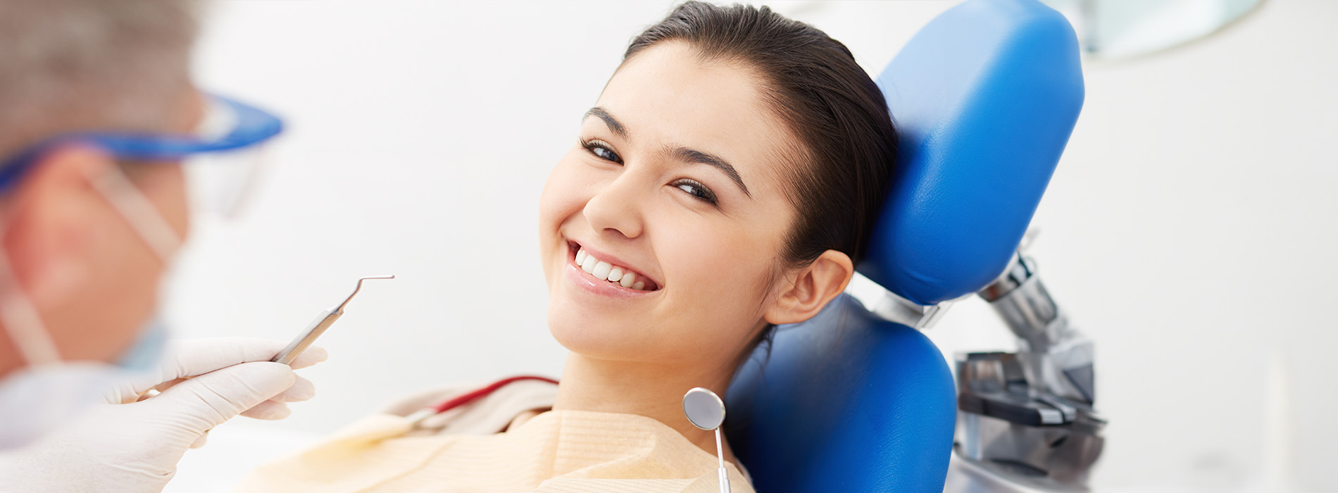 The image shows a dental setting with a smiling woman sitting in the dentist s chair, receiving care from a professional who is wearing a blue face mask and a white coat.