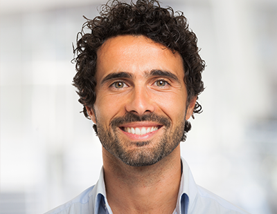 A smiling man with a beard, wearing a blue shirt and standing in front of a building.