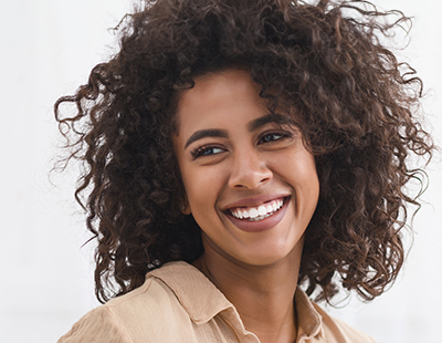 The image shows a smiling woman with curly hair and a bright smile, wearing a tan top.