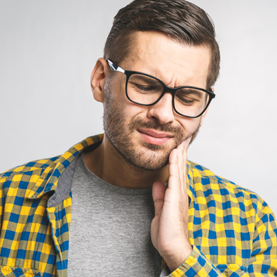 The image shows a man with glasses, wearing a yellow plaid shirt and holding his face with one hand, possibly in pain or discomfort.
