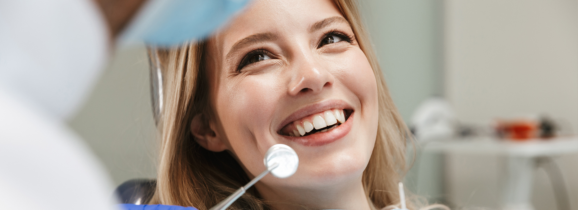 A woman in a dental office, smiling at the camera, with a dentist in the background.