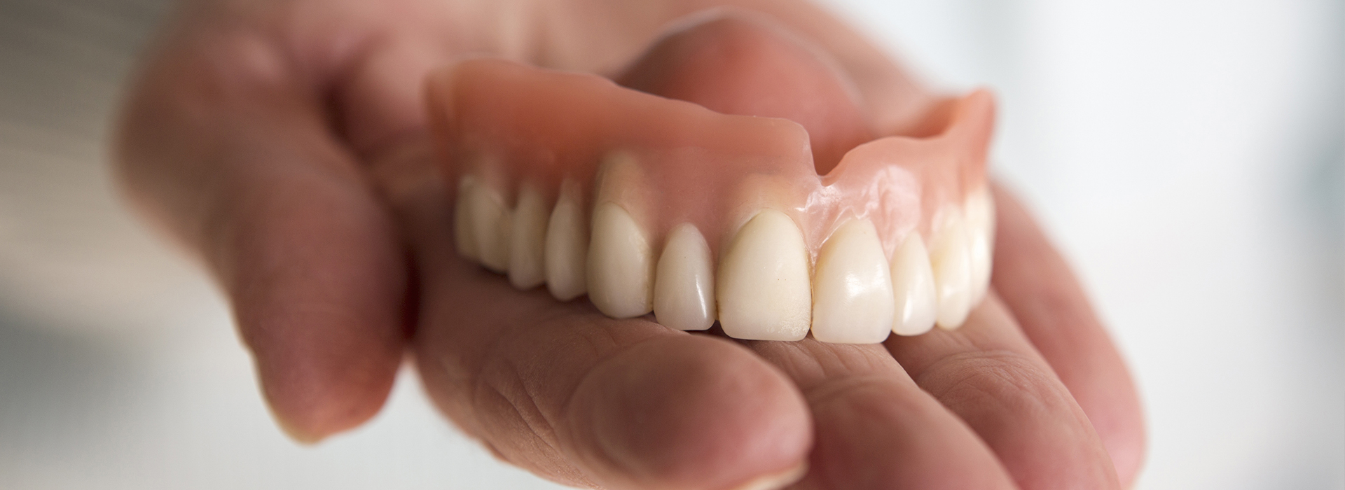 An image featuring a person s hand holding a set of dentures against a blurred background.