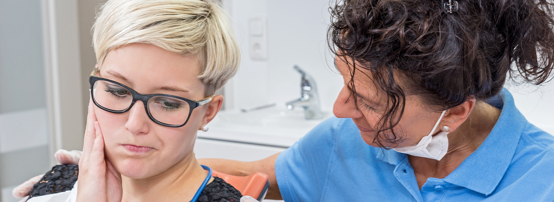 A woman in a blue shirt and glasses is sitting in a dental chair, receiving dental care from a female dentist wearing a white coat.