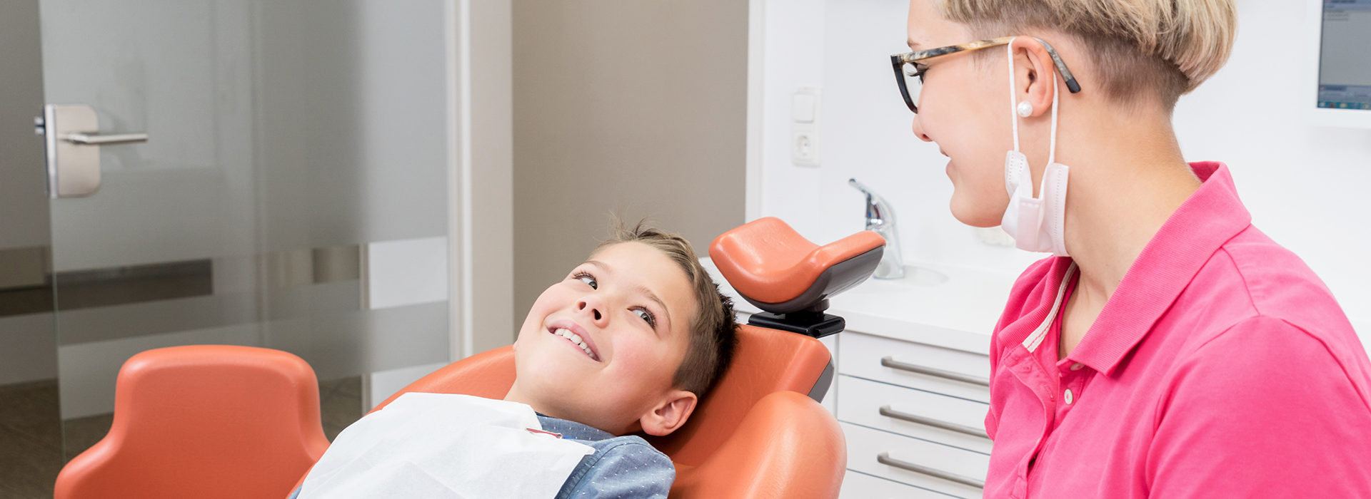 A young child is seated in a dental chair, receiving dental care from an adult wearing a pink shirt.