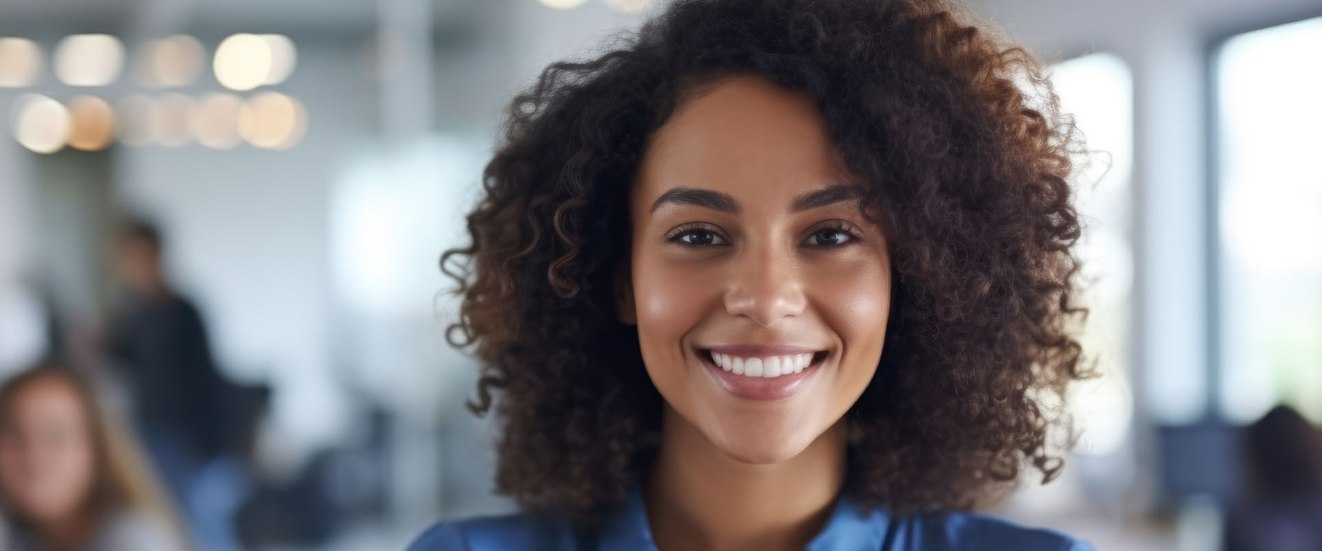 The image shows a person with curly hair, smiling at the camera, wearing a professional outfit in an office setting.