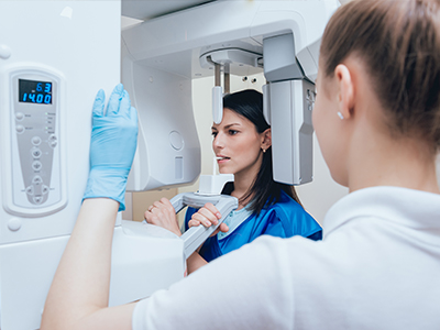 A woman in a blue uniform stands next to a large, modern 3D scanner, with another person who appears to be an operator or technician in the background.