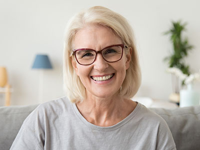 A smiling woman with short blonde hair, wearing glasses and a light-colored top, seated indoors in front of a homey background.