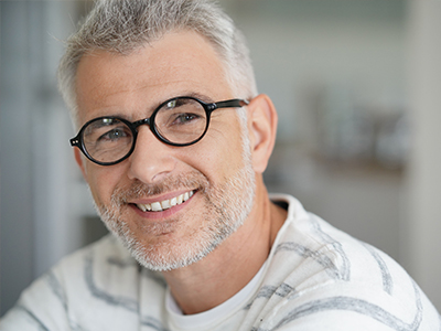 The image features a man with glasses, smiling at the camera. He has short hair and appears to be middle-aged. The background is blurred but suggests an indoor setting, possibly an office or home environment.