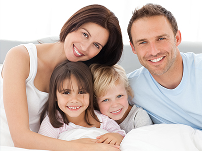 A family of four, including two adults and two children, smiling together in a cozy bedroom setting.