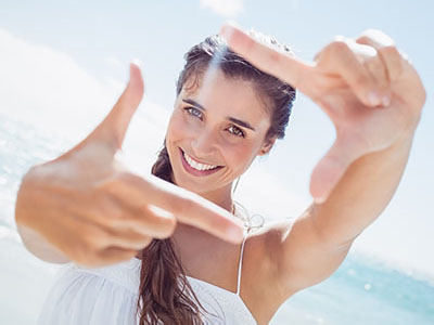 A young woman with long hair, smiling at the camera, holding her hand up to create a frame around herself.